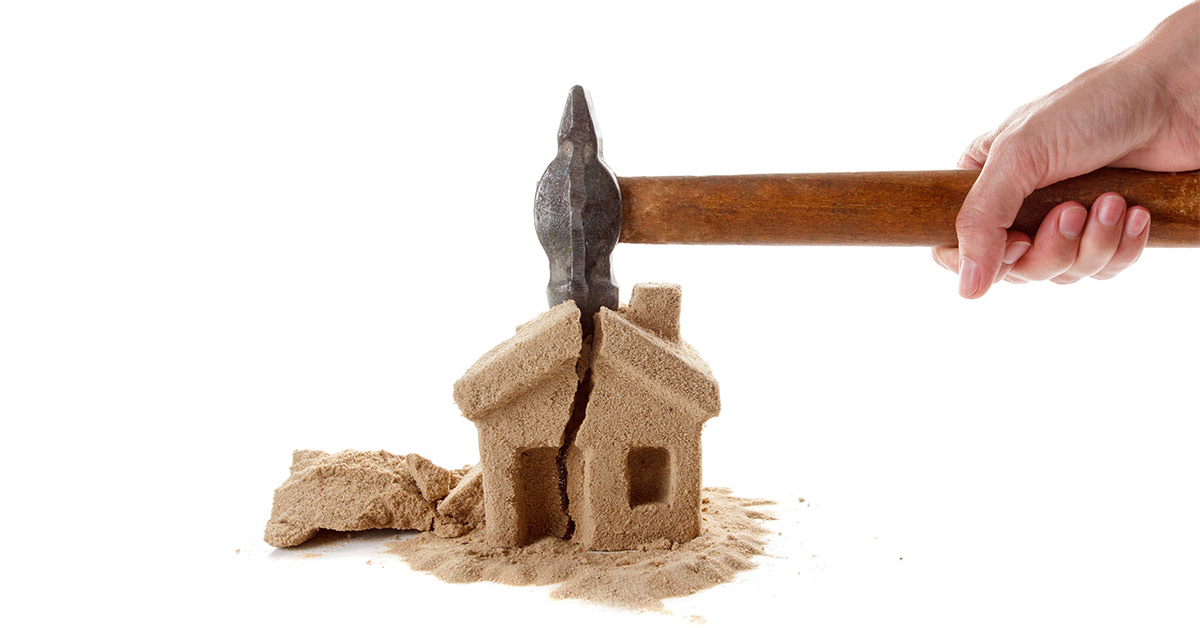 Eine Hand schlägt mit einem Hammer auf ein Haus aus Sand und zerstört es | Notverkauf Immobilie